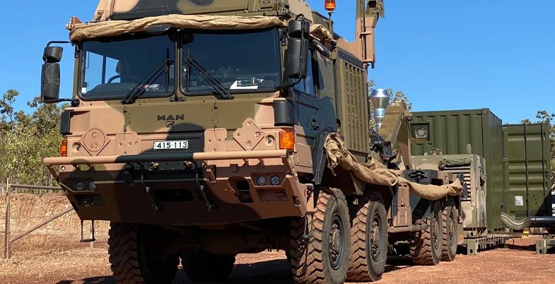 Camouflaged military truck carrying a deployable shelter on a red-dirt track in the Australian bush